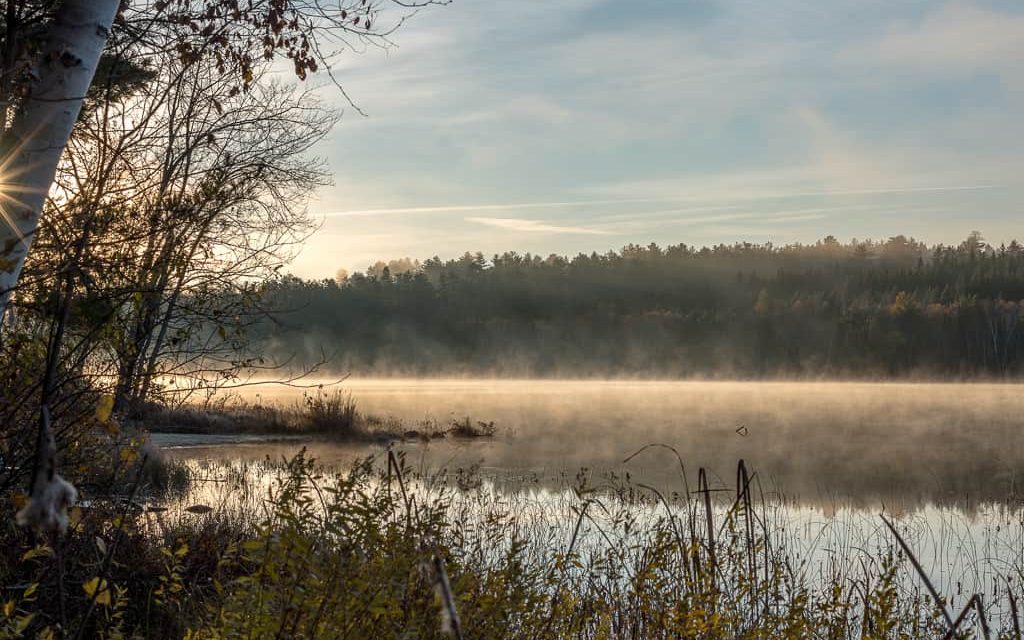 The morning sun rises, revealing fog and a chair sits on a partially submerged dock beside Three Mile Lake.